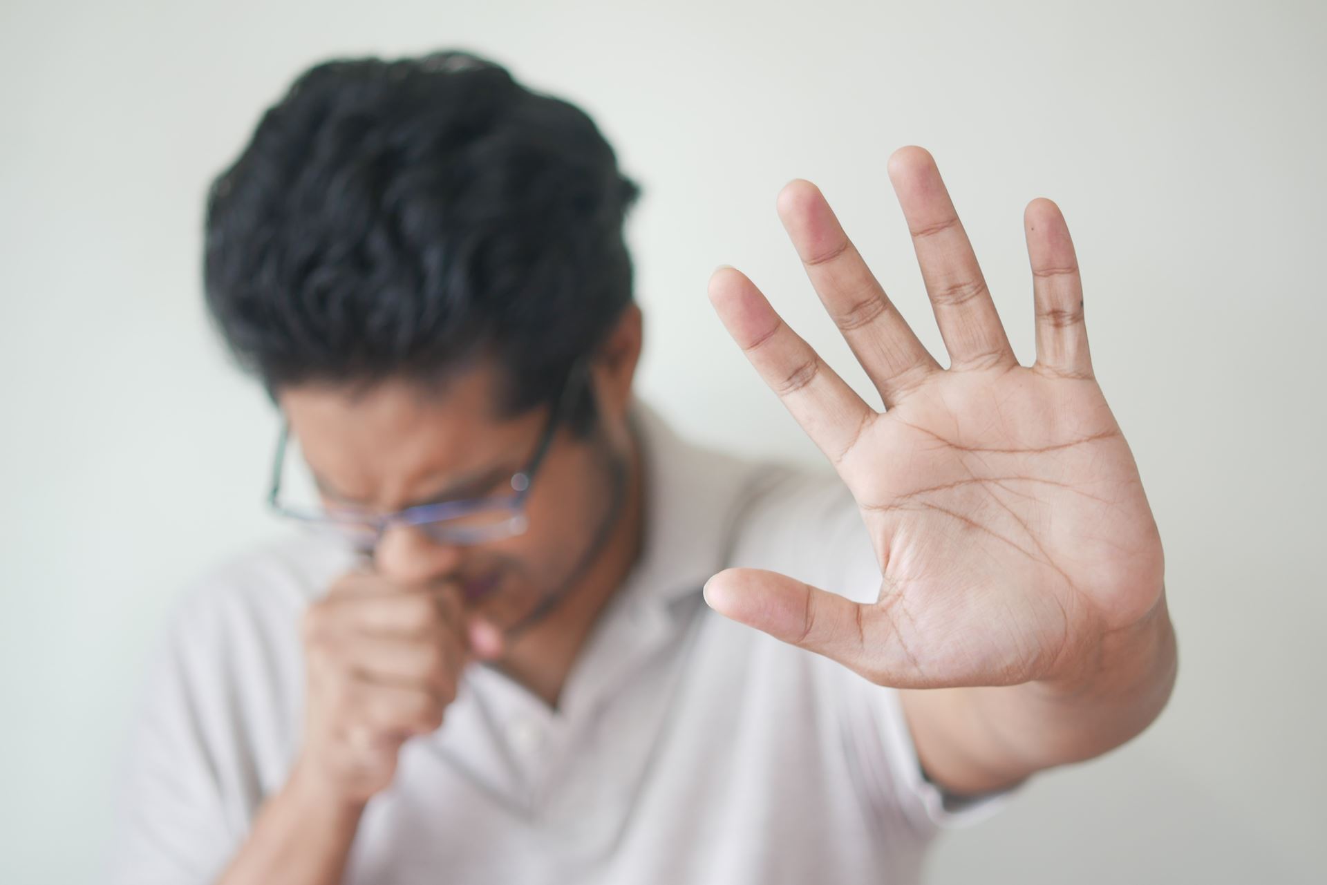 a man coughing, covering his mouth with his right hand and stretching the open palm of his left hand in a gesture to keep away