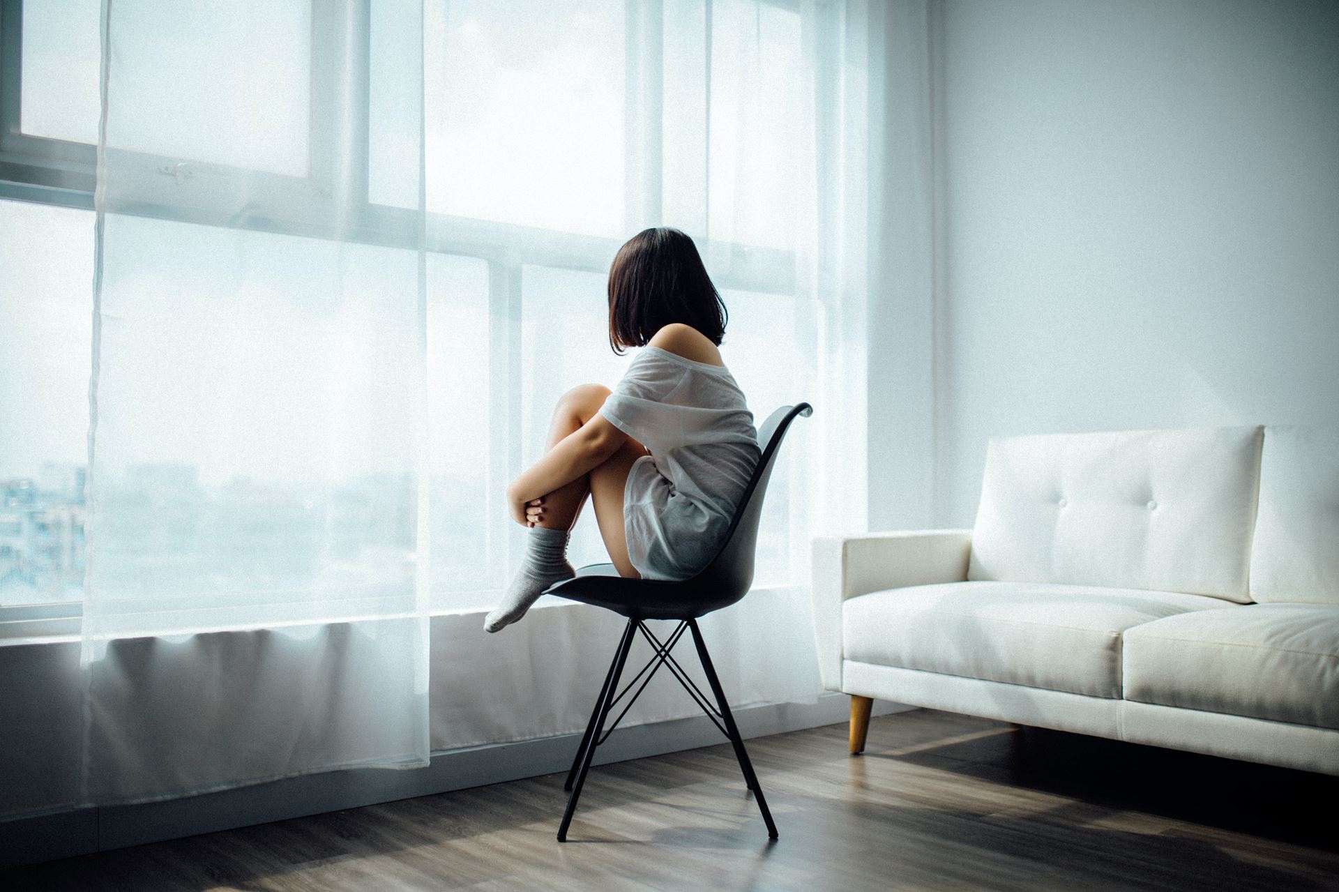 a woman sat on a chair, hunched up, her arms around her legs looking out a window